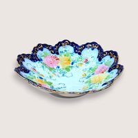 Scalloped porcelain bowl with a deep cobalt blue rim and intricate gold filigree. The interior features hand-painted pink and yellow floral bouquets with delicate green vines on a white ground.