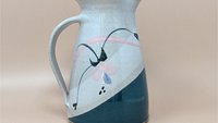 Speckled cream earthenware pitcher with a wide flared rim and sturdy handle. Features a hand-painted abstract floral design in teal, pink, and indigo with a diagonal teal color-block base.