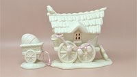 Two-piece Dept. 56 Snowbunnies set featuring a large cottage-style wagon and a small egg cart. Matte white bisque porcelain with soft pink accents and hanging Easter egg garlands.