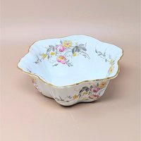 A scalloped white porcelain bowl with a gold gilt rim. Features pink and yellow rose bouquets with grey-green leaves draped across the interior and exterior. Delicate, fluted silhouette with a glossy finish on a neutral tan background.