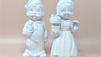 Pair of 7-inch ceramic figurines of an elderly man in striped pajamas with a candle and a woman in a nightgown holding a gift. White pearlescent luster glaze with soft blue accents.