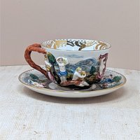 Italian Capodimonte porcelain cup and saucer set. Features high-relief, hand-painted classical figures in a woodland landscape. Intricate gold gilt interior accents and a rustic coral-style handle. Cup 2.5", Saucer 6".