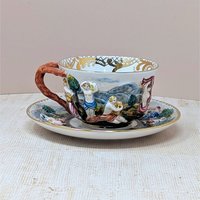 Italian Capodimonte porcelain cup and saucer set. Features high-relief, hand-painted classical figures in a woodland landscape. Intricate gold gilt interior accents and a rustic coral-style handle. Cup 2.5", Saucer 6".