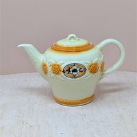 A vintage Porcelier cream-colored teapot with a raised, textured sunflower and rope border in amber-brown tones. A central black and white floral medallion sits within an oval frame. The teapot has a classic rounded shape and matching lid, set against a neutral, warm background.