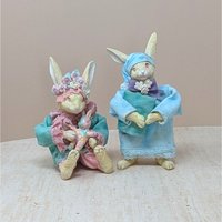 Two whimsical vintage rabbit figurines dressed in pajamas. One wears a blue nightgown and sleeping cap holding a book; the other wears a teal coat and flower crown, holding a small pink rabbit doll.