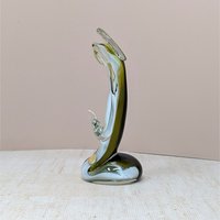 A handmade art glass figurine with a smooth, fluid silhouette. The piece features a vibrant emerald green core encased in a clear, glossy glass exterior. Its organic, elongated form tapers elegantly, reflecting light across its polished surface.
