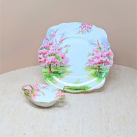 Two-piece Royal Albert Blossom Time bone china set. Includes a 9.75-inch square handled cake plate and a 5.5-inch oval handled sweet meat dish. Features pink blossom trees and gold-trimmed scalloped edges.