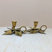 A pair of solid brass chamberstick candle holders. Each base is shaped like two large, overlapping maple leaves with detailed veining. Features a finger loop handle and a single tulip-style taper cup.