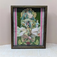 A vertical shadow box frame containing a Victorian-style paper collage. Features a cherub, floral lithographs, and a large pink velvet bow against a forest green velvet background. Housed in a dark wood-grain frame.