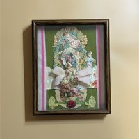 A vertical shadow box frame containing a Victorian-style paper collage. Features a cherub, floral lithographs, and a large pink velvet bow against a forest green velvet background. Housed in a dark wood-grain frame.