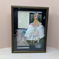 Dark wood shadow box featuring a 3D collage of a Victorian woman in a white lace dress. Black background with a frosted perfume bottle, dried botanicals, and handwritten yellowed paper. Rich, layered multimedia textures.