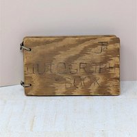 A small, rectangular autograph book with a natural wood grain cover. The words "Autograph Book" are hand-carved into the wood. Two metal rings bind the pages on the left. Rustic, weathered texture with a warm honey-oak finish.