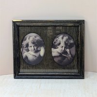 A dark wood frame with two oval cutouts. Inside are sepia-toned prints of a young child with curly hair and wings, depicting "Angel Awake" looking at the viewer and "Angel Asleep" with eyes closed. Rustic wood grain texture.