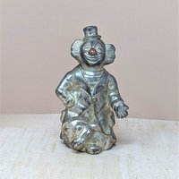A vertical silver-toned metal bank shaped like a circus clown in a top hat and suit. Features an aged metallic patina with a small red detail on the nose. Sculptural, heavy-duty texture with a slot on the reverse.