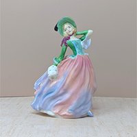 A vertical porcelain figurine of a woman in a wind-swept gown. She wears a vibrant green bodice, a wide-brimmed hat, and a voluminous skirt in blended shades of pink, peach, and lavender. Glossy, fine-bone china texture.