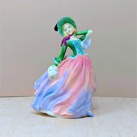 A vertical porcelain figurine of a woman in a wind-swept gown. She wears a vibrant green bodice, a wide-brimmed hat, and a voluminous skirt in blended shades of pink, peach, and lavender. Glossy, fine-bone china texture.