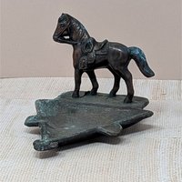 A vertical cast metal ashtray featuring a saddled horse figurine standing on a triangular base. The metal has a dark bronze patina with heavy green verdigris oxidation throughout. Sturdy, industrial texture with period-correct relief details.
