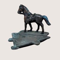 A vertical cast metal ashtray featuring a saddled horse figurine standing on a triangular base. The metal has a dark bronze patina with heavy green verdigris oxidation throughout. Sturdy, industrial texture with period-correct relief details.