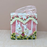 A square ceramic vase shaped like a gift bag with pink ribbon handles. The front features a high-relief scene of two white bunnies in a garden with pink roses and yellow lace detailing. Glossy vitrified texture.