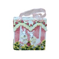A square ceramic vase shaped like a gift bag with pink ribbon handles. The front features a high-relief scene of two white bunnies in a garden with pink roses and yellow lace detailing. Glossy vitrified texture.