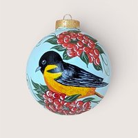 A spherical glass ornament featuring a hand-painted Baltimore Oriole bird with a black head and vibrant orange breast. Surrounded by clusters of bright red berries and green leaves. Topped with a classic gold metal cap.