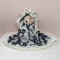 A vertical 12-inch handcrafted teddy bear with tan fur and movable limbs. Wearing an elaborate Victorian-style dark floral dress with voluminous cream lace trim and a matching oversized lace bonnet.