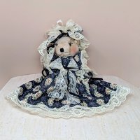 A vertical 12-inch handcrafted teddy bear with tan fur and movable limbs. Wearing an elaborate Victorian-style dark floral dress with voluminous cream lace trim and a matching oversized lace bonnet.