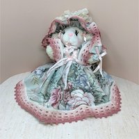 A seated plush lamb in a wide, floral-patterned sage green and rose pink dress. The lamb features long eyelashes, a pink nose, and an oversized matching lace bonnet. The intricate pink lace trim on the hem and bonnet adds a grounded, romantic texture.