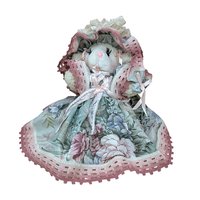 A seated plush lamb in a wide, floral-patterned sage green and rose pink dress. The lamb features long eyelashes, a pink nose, and an oversized matching lace bonnet. The intricate pink lace trim on the hem and bonnet adds a grounded, romantic texture.