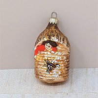 A vertical 4.5-inch vintage blown glass ornament shaped like a beehive. A hand-painted baby with a red suit peeks from the top, and a large brown bee is featured on the front. Silvered glass with gold glitter accents.