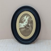 An oval vintage sepia-toned print titled "Cupid Asleep" by M.B. Parkinson. It depicts a young child with curls leaning over a bow. The print is housed in a thick, matte black wooden oval frame. 8.5 by 6.75 inches.A vintage sepia-toned oval print of M.B. Parkinson's "Cupid Asleep" featuring a young child with wings holding a bow. Housed in a black oval wooden frame with visible surface scrapes and aging.