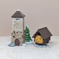 A set of two hand-painted porcelain figurines including a tall stone silo with a brown roof, a snowy evergreen tree, and a small wooden hayshed filled with golden hay. Realistic textures and snow-dusted bases.