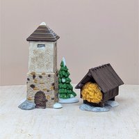 A set of two hand-painted porcelain figurines including a tall stone silo with a brown roof, a snowy evergreen tree, and a small wooden hayshed filled with golden hay. Realistic textures and snow-dusted bases.