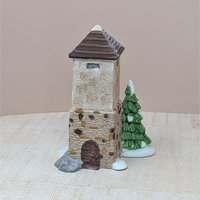 A set of two hand-painted porcelain figurines including a tall stone silo with a brown roof, a snowy evergreen tree, and a small wooden hayshed filled with golden hay. Realistic textures and snow-dusted bases.