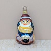 A hand-blown glass ornament of a cheerful gnome with a white beard and red hat, sitting on a brown log and playing a gold accordion. Features a gold star-shaped hanging cap. 4.25 inches tall