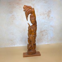 A vintage mid-century hand carved wooden statue of a Balinese woman on a tan textured background. Standing twenty eight inches tall, the highly detailed timber figure poses gracefully beneath an ornate, stylized palm frond canopy.