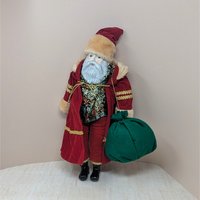 An 18-inch Santa Claus figure standing on a white base. He wears a deep red velvet robe with gold braided trim and a fur-trimmed hood, holding a green velvet toy sack and featuring a detailed ceramic face.