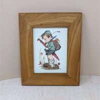 A completed and framed cross-stitch of a young boy in traditional Alpine clothes holding a large red umbrella, known as the Hummel "It Looks Like Rain" design. 11 by 9 inches.