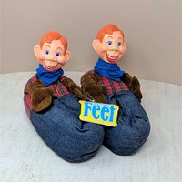 A pair of vintage child-sized slippers featuring the plastic head of Howdy Doody with a red plaid shirt and denim-look base. Original yellow "Feet" tag attached.