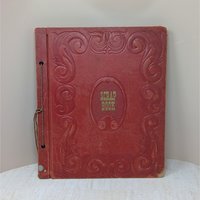 A large, vintage red leatherette scrapbook with an ornate embossed border and "Scrapbook" in gold lettering at the center. The cover shows significant age-related wear and is secured by a brown cord binding.