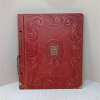 A large, vintage red leatherette scrapbook with an ornate embossed border and "Scrapbook" in gold lettering at the center. The cover shows significant age-related wear and is secured by a brown cord binding.