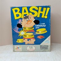 The original 1965 box for the Milton Bradley game "BASH!" featuring a bright blue background and a cartoon man being "bashed" apart. The box shows vintage shelf wear but remains structurally sound with vibrant yellow lettering.