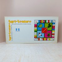A long white rectangular game box for Agri-Venture. The text is in a retro orange and yellow serif font. A colorful graphic of the square game board is on the right, featuring illustrations of a cow, corn, and trees.