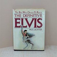 A large hardcover book titled The Boy Who Dared To Rock: The Definitive Elvis. The white dust jacket features an illustration of Elvis Presley in a pink jacket with a guitar. The jacket shows visible edge wear and small tears.
