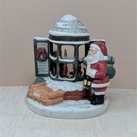 A painted ceramic music box featuring santa standing by a snowy bay window. A small child is visible inside the house through the panes. The colors are classic red, evergreen, and snowy white with a matte finish.