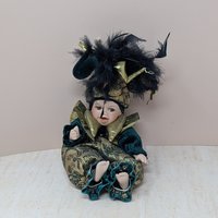 A 12-inch porcelain baby doll dressed as a jester in emerald green velvet and gold brocade, featuring a large feathered jester hat and gold metallic ruff.