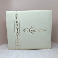 A large, square vintage photo album in cream faux leather. The word Memories is printed in a gold cursive script on the front. A decorative vertical border of gold filigree crosses runs down the left side.