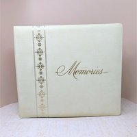 A large, square vintage photo album in cream faux leather. The word Memories is printed in a gold cursive script on the front. A decorative vertical border of gold filigree crosses runs down the left side.