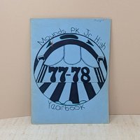 A vintage softcover yearbook featuring a light blue cover with a hand-drawn circular graphic in black ink. The center shows the bold numbers 77-78 under a theatrical or arched motif. A name is written in small ink at the top right.