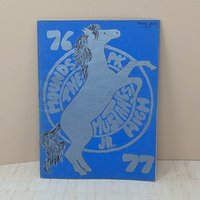 A vintage softcover yearbook with a bold blue cover. It features a large, silver-ink illustration of a rearing mustang horse centered over a circular school seal. The year 76-77 is printed in large block numbers.